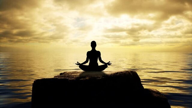 A female silhouette sitting on a rock at the ocean's edge, meditating in the lotus position in-front of a beautiful golden sunset.