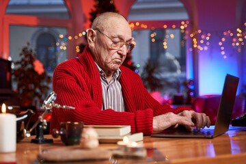 Fairy and lovely room decorated with xmas tree and lights and an old man typing on modern laptop sitting at table.
