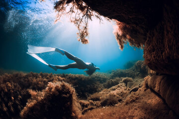 Young woman freediver girl with fins glides near rock cave with sun rays. Freediving underwater in...