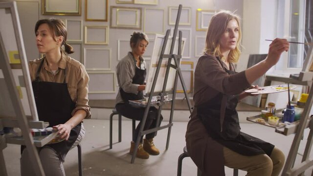 Full shot of three young concentrated women wearing shirts and aprons sitting and painting on canvas at easel attending arts masterclass in bright modern studio