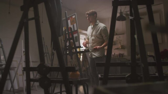 Medium shot of young male painter entering dark art studio, approaching to easel with self-made abstract painting, making finishing strokes enjoying process