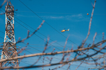 Helicóptero amarillo visto a través de ramas en la distancia. Volando entre torres eléctricas de alta tensión.