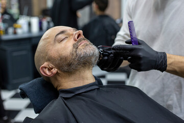 Portrait of client during beard grooming in barber shop. Barber forms the shape of customer's beard with a trimmer. Selective focus