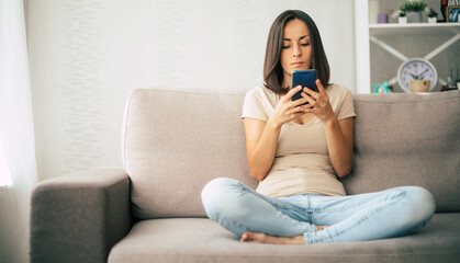 Fototapeta premium Young modern confident smiling woman is working or relaxing at home and sitting on the couch while using her smartphone