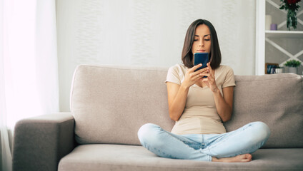 Fototapeta premium Young modern confident smiling woman is working or relaxing at home and sitting on the couch while using her smartphone