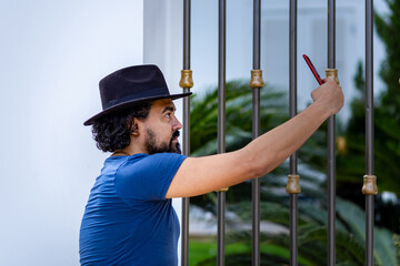 Homem com barba e chapéu, fazendo selfie com o uso de um celular.