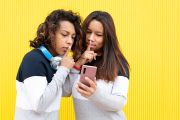 Portrait of two brown girls on a yellow background. Both are making the gesture of asking for silence while looking at a smart phone. Space for text. 