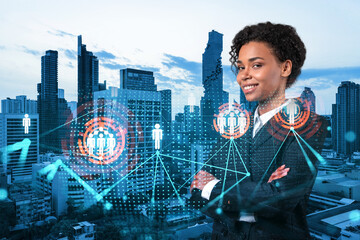 Smiling black woman HR director at international company is thinking about recruitment of highly qualified specialists. Women in business concept. Social media hologram icons over Bangkok.
