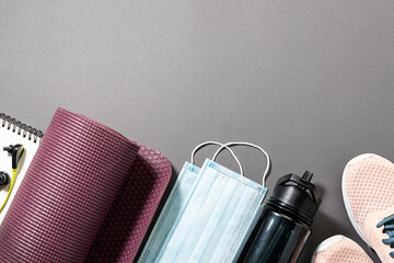 Fitness accessories for quarantine time top view. Notebook, headphones, water bottle, sneakers, yoga mat and protective masks on gray background.