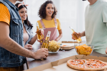 Potato chips and takeaway pizza near hispanic friends with beer at home on blurred background