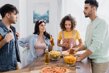 Hispanic friends talking near pizza, chips and bottles of beer in kitchen