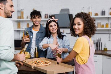 Smiling hispanic woman looking at camera near friends opening beer and pizza in kitchen