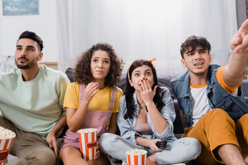 worried hispanic man pointing with finger while watching movie near nervous friends with popcorn, blurred foreground