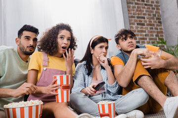 shocked hispanic friends with popcorn buckets watching tv at home