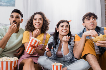 cheerful hispanic friends eating popcorn and chips while watching tv