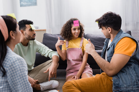 hispanic friends showing thumbs up while sitting on floor and playing guess who game