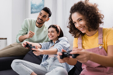Excited Hispanic man holding clenched fist near cheerful friends gaming with joysticks, blurred...