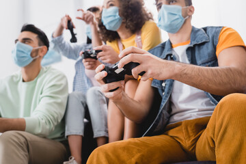 Obraz premium Gamepad in hands of hispanic man in medical mask playing video game with friends on blurred background