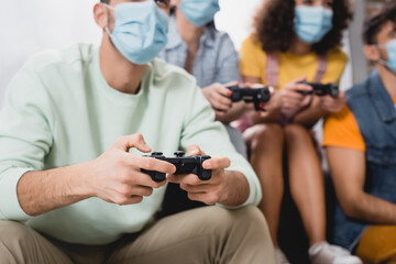 Cropped view of man in medical mask holding joystick near friends on blurred background at home