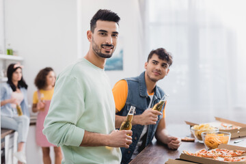 Smiling hispanic man holding bottle of beer near friends, pizza and chips on blurred background