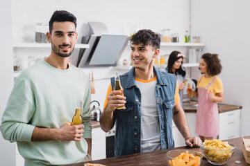 Positive hispanic men holding bottles of beer near potato chips and friends on blurred background