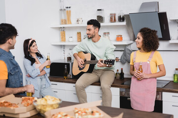 Smiling hispanic friends with beer looking at man playing acoustic guitar near food on blurred foreground