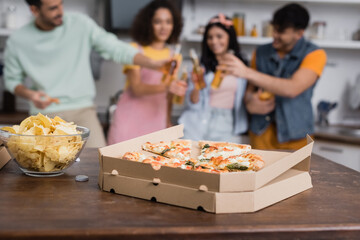 Takeaway pizza and potato chips on table near friends with beer on blurred background in kitchen