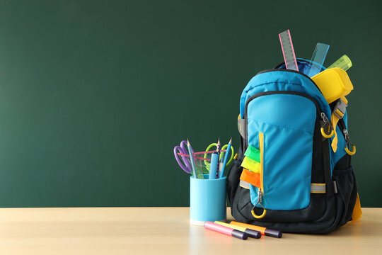 Stylish backpack and different school stationery on wooden table near green chalkboard, space for text. Back to school