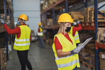 Young woman at work in warehouse with digital tablet scanner while wearing protective face mask for coronavirus - People working staff in background