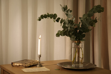 Beautiful eucalyptus branches, old book and holder with burning candle on wooden table indoors. Interior element