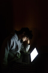 Young man with grey hoodie illuminated by a white laptop screen, touching his face with his hand...
