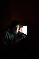 Young man with face mask and grey hoodie illuminated by a white laptop screen, looking at camera with surprise expression. Dark orange wall in the background