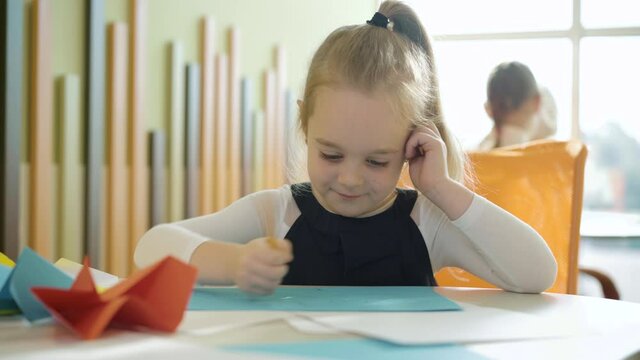 Cute happy girl drawing picture with classmates sitting at the background. Portrait of positive pretty Caucasian schoolgirl on art lesson in school. Talent and creativity concept.