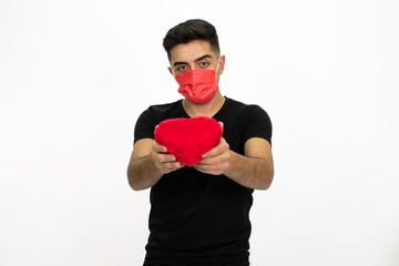 Young male model wearing a red medical face mask. He is holding a gift box in the shape of a heart. He is wearing a black shirt. Isolated image and white background.