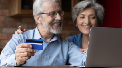 Focus on plastic credit bank card in old wrinkled male hands. Happy middle aged family couple enjoying purchasing goods in internet store, involved in online shopping using computer app at home.