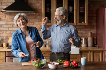 Overjoyed emotional middle aged senior married family couple having fun preparing food together in kitchen, having playful mood. Sincere happy old husband and wife laughing, cooking meal at home.