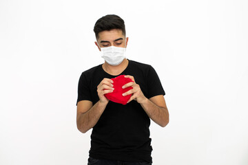 Young male model wearing a medical face mask. He is holding a gift box in the shape of a heart. He is wearing a black shirt. Isolated image and white background.