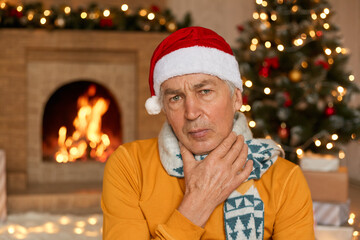 Sick man in orange sweater, scarf and Christmas hat suffering from sore throat, keeping hand on neck, posing on background of fireplace and fir tree, spends new year eve ant home.