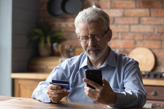 Focused mature retired man holding bank credit card, entering payment information in mobile shopping application, purchasing goods or services in online store, old people and modern tech concept.