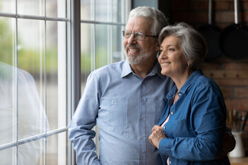 Happy bonding loving middle aged senior retired couple standing near window, looking in distance, recollecting good memories or planning common future, enjoying peaceful moment together at home.
