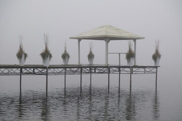 Romantic moody scenery with wooden pier with white gazebo by lake on calm misty day