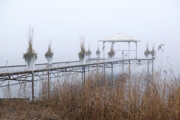 Fototapeta na wymiar Romantic moody scenery with wooden pier with white gazebo by lake on calm misty day