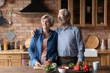 Happy middle aged family couple dreaming, distracted from preparing food together in kitchen. Smiling sincere loving old wife and husband looking in distance, enjoying peaceful weekend morning.