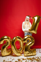 Xmas smiling young girl in santa hat with golden balloons in 2021 shape on red background for christmas holiday design. High quality photo