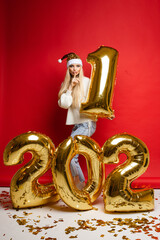 Gold xmas balloons in 2021 shape, young girl in santa hat shows to be quiet on red studio background with copy space. High quality photo