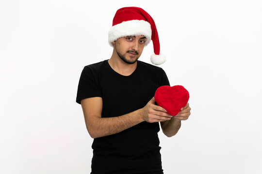 Young man wearing a Santa hat. He is holding a gift box in the shape of a heart. He is wearing a black shirt. Isolated image white background.