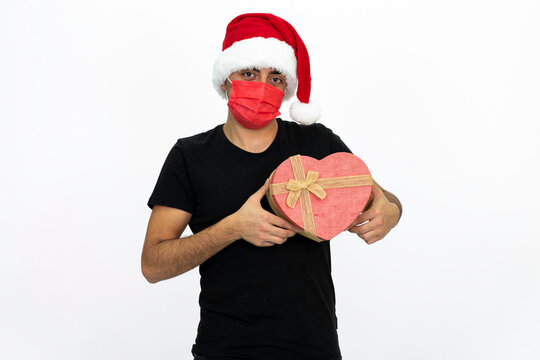 Young man wearing a Santa hat. He has a red medical face mask. She is holding a heart-shaped box in her hand. He is wearing a black shirt. Isolated image white background.