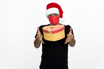 Young man wearing a Santa hat. He has a red medical face mask. She is holding a heart-shaped box in her hand. He is wearing a black shirt. Isolated image white background.