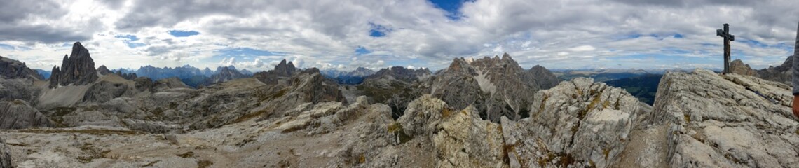 360-degree view from Italian Dolomites. There is a wooden cross on a mountain peak. In the back there are endless mountain chains. Raw and unspoiled landscape. Few clouds above. Sharp and steep slopes