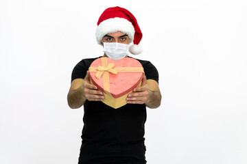 Fototapeta na wymiar Young man wearing a Santa hat. She is holding a heart-shaped box in her hand, wearing a medical face mask.